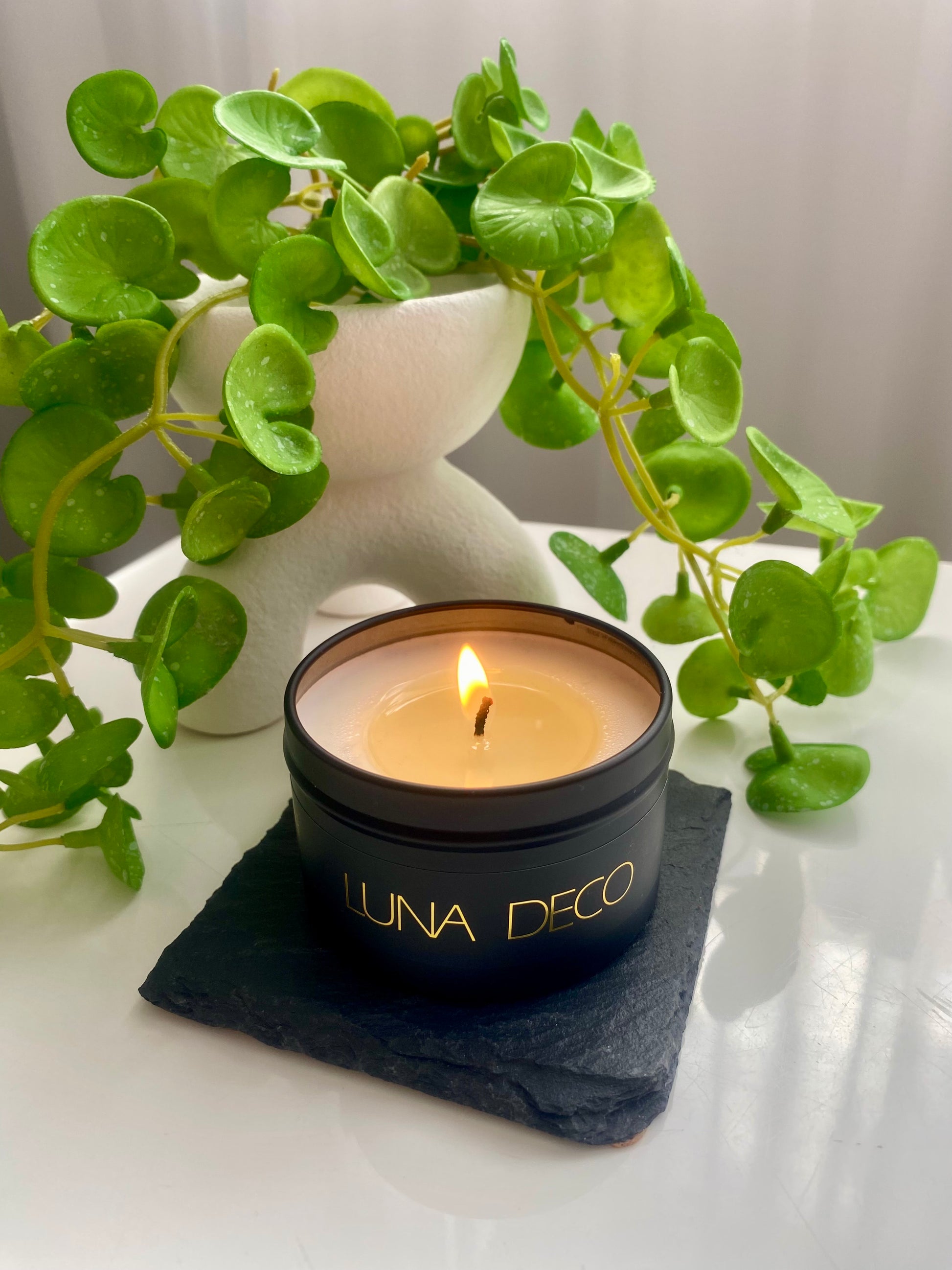 Luna Deco candle with greenery in the background.
Matte black travel tin candle,  perfect for travel, small spaces or thoughtful gifting.
Minimalist, timeless, elegant design, artisan-made candles. 
Luna Deco. 